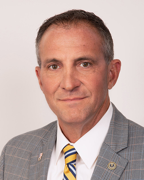 The image is a portrait-style photograph of an older adult male wearing a suit with a tie, standing against a plain background. He has short hair, appears to be middle-aged, and is smiling slightly at the camera.