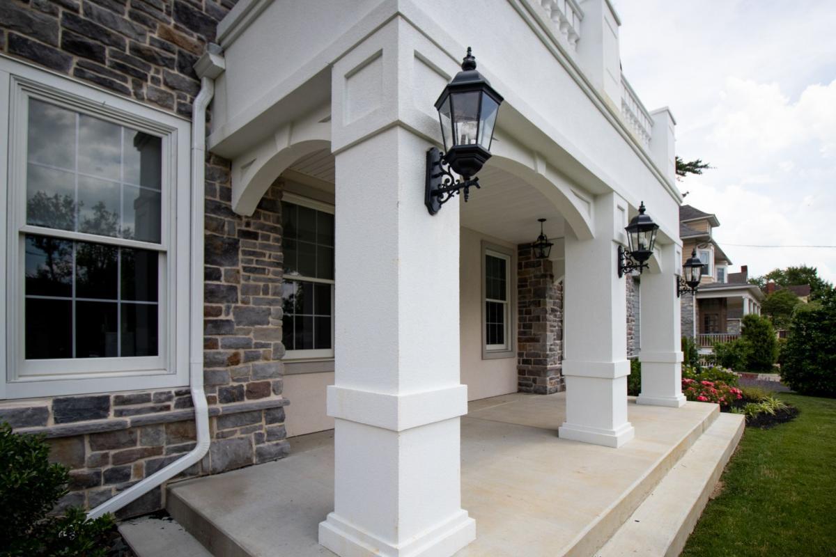 The image shows a white porch with pillars, flanked by two black lampposts, leading up to a front door of a house, featuring a brick facade and a large window above the entrance.