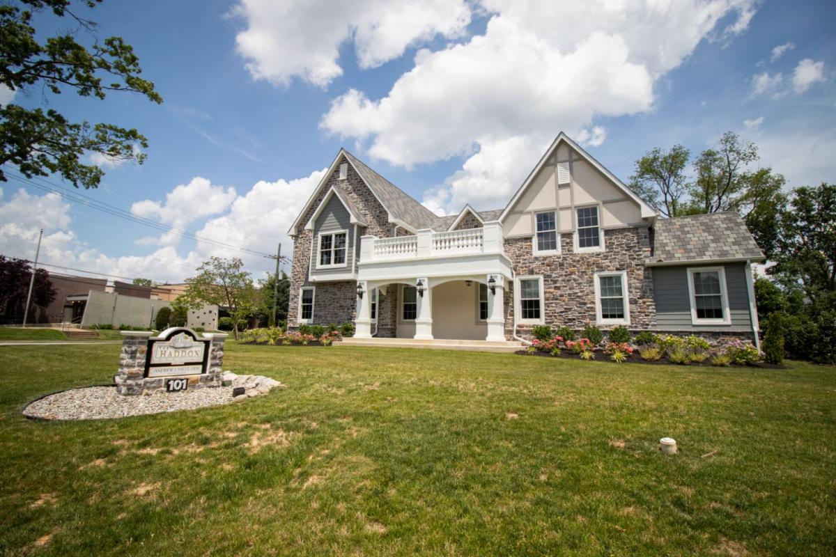 The image shows a two-story residential house with a prominent front porch, set against a clear sky with some clouds, and surrounded by a well-maintained lawn and landscaping.