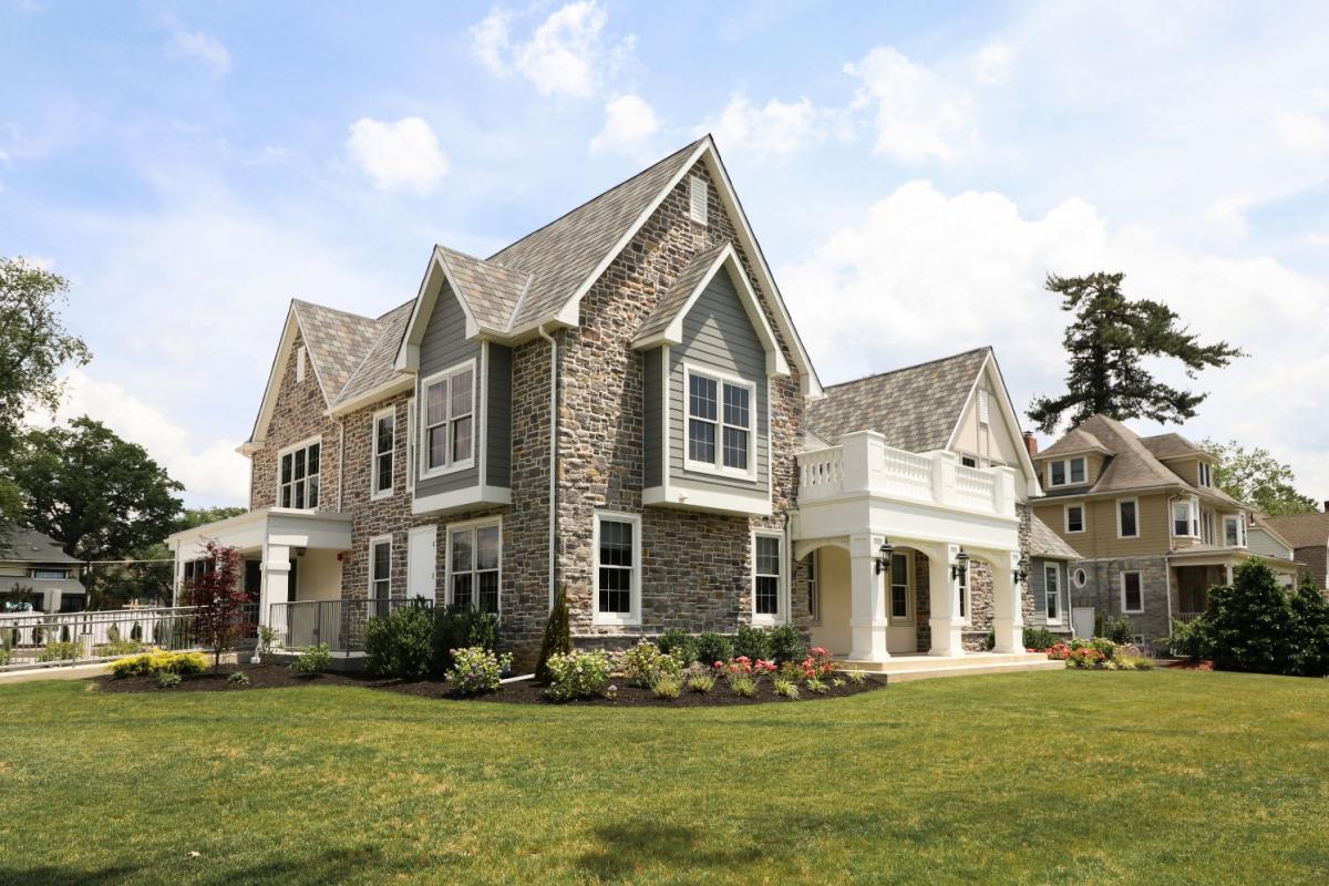 The image depicts a two-story residential house with a combination of stone and brick exterior walls, a prominent front porch with white railings, and a manicured lawn with a few trees and shrubs.