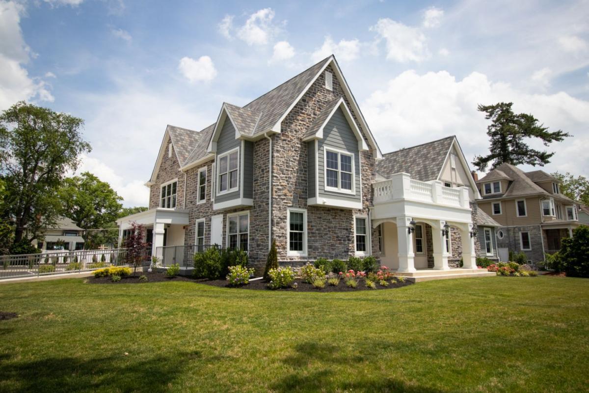 The image depicts a two-story residential house with a stone facade, white trim around windows and doors, and a combination of brick and siding exterior. The house features a prominent front porch with columns, a well-manicured lawn, and a clear sky in the background.