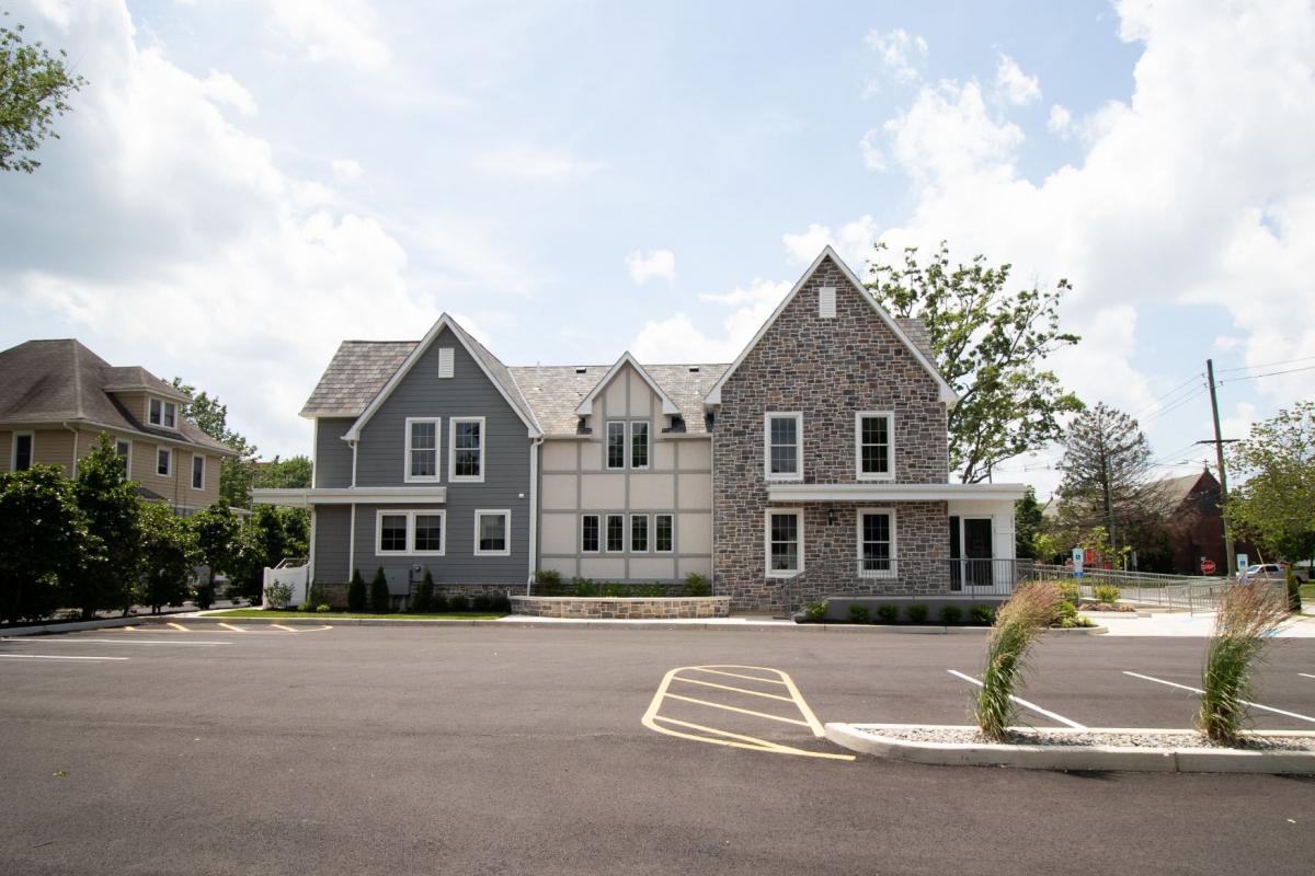 The image depicts a two-story residential house with a gray exterior, a white trim around the windows and doors, and a stone facade on the front. There are multiple windows visible on both levels of the house, with a prominent bay window on the second floor. A car is parked in the driveway, which leads to a garage door. The house has a manicured lawn and is situated in a suburban neighborhood with other similar homes nearby.