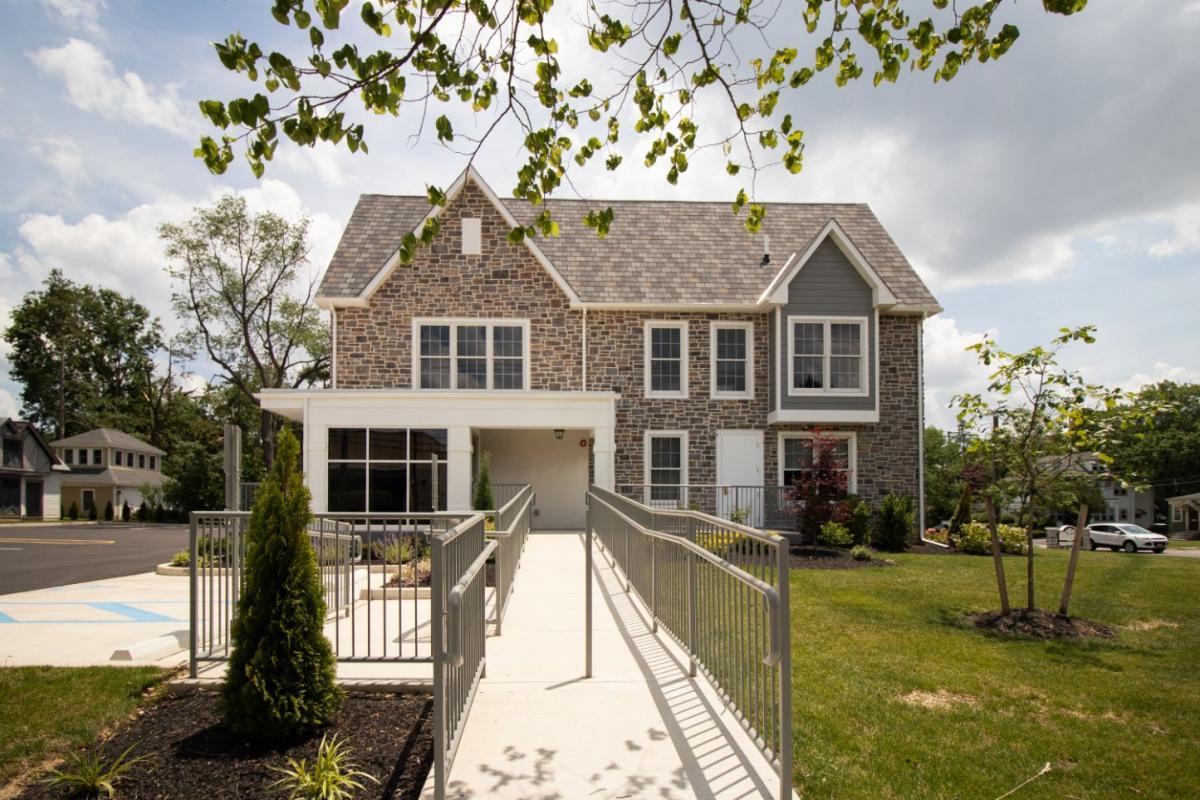 The image depicts a two-story house with a prominent front porch and a brick facade, under a clear sky with some clouds.