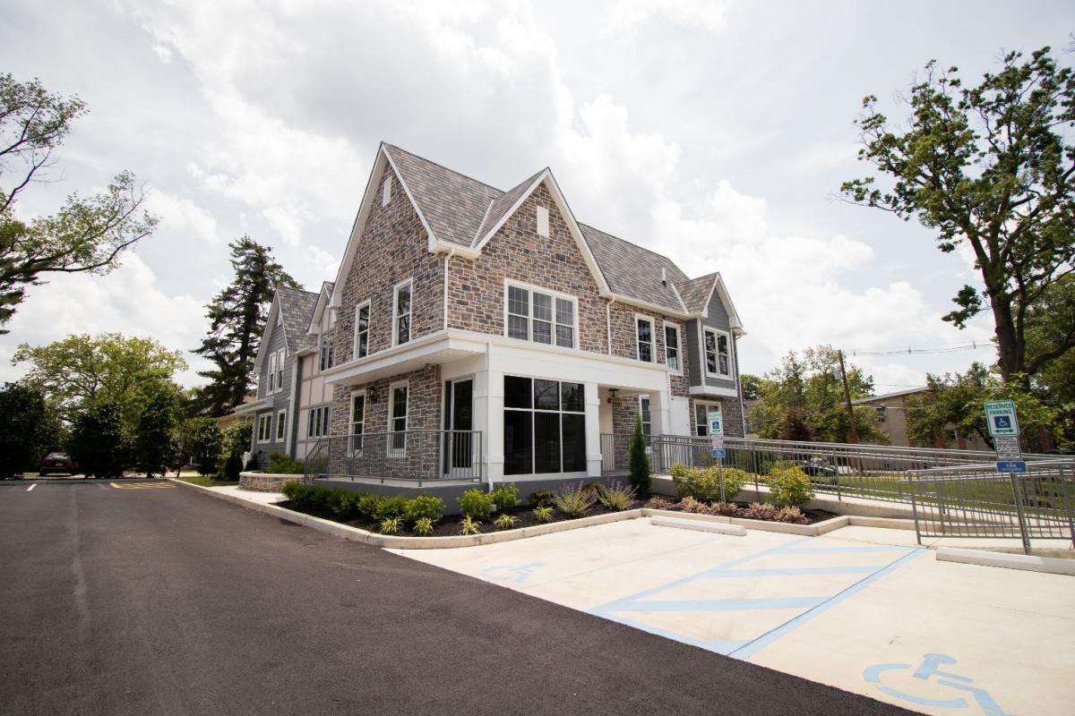 The image shows a two-story building with a prominent front entrance, featuring a large window flanked by white columns, a stone facade, and a covered porch with a railing. The architecture suggests a formal and possibly institutional design.