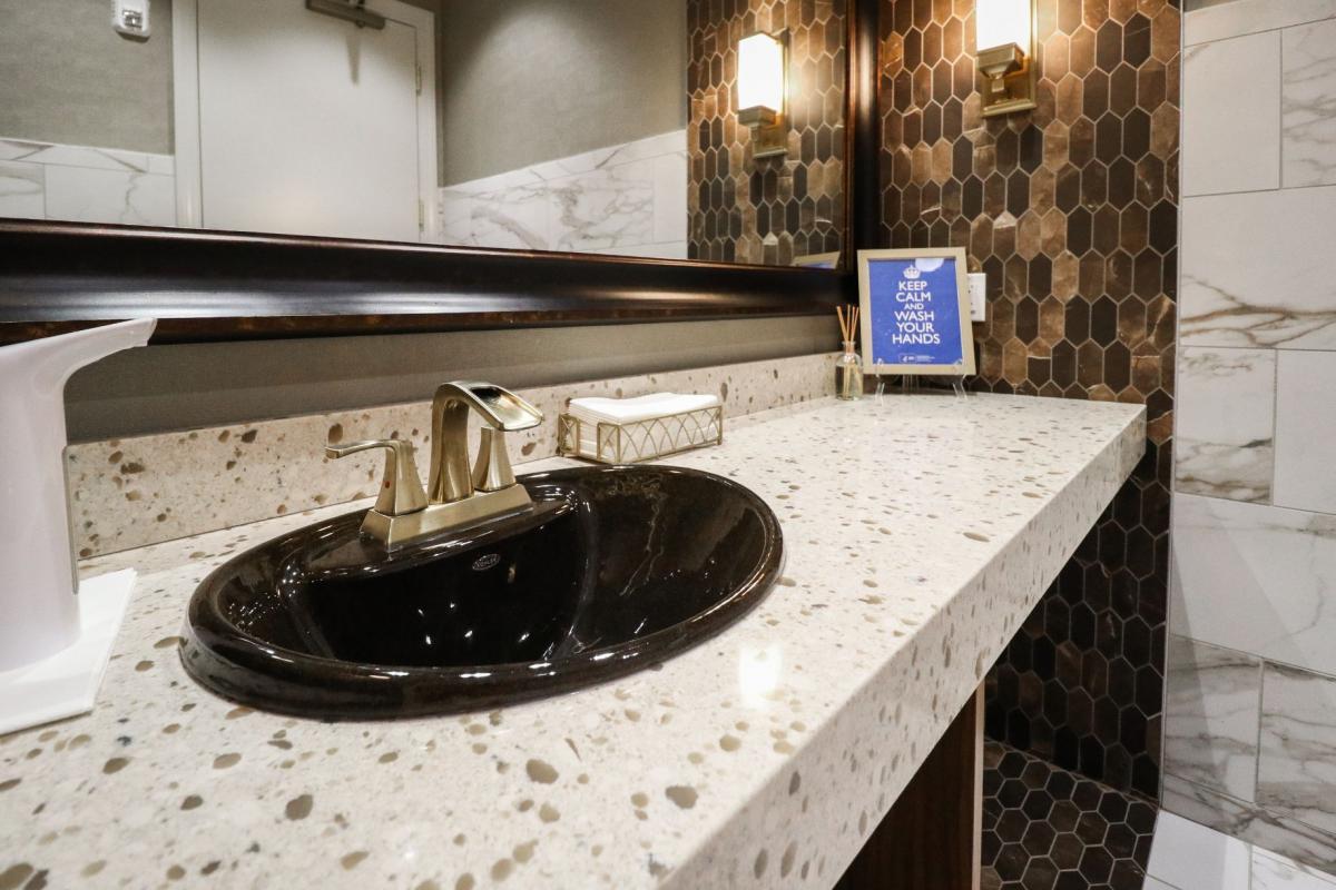 The image shows a modern bathroom interior with a marble countertop, a black sink, gold fixtures, and a white tile backsplash.