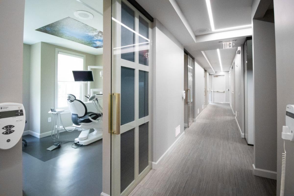 The image depicts a modern, well-lit hallway with a sleek design, featuring a combination of gray walls, dark flooring, and contemporary furnishings such as a reception desk, seating area, and a restroom. There are also medical equipment visible in the background, suggesting that this space is within a healthcare facility.