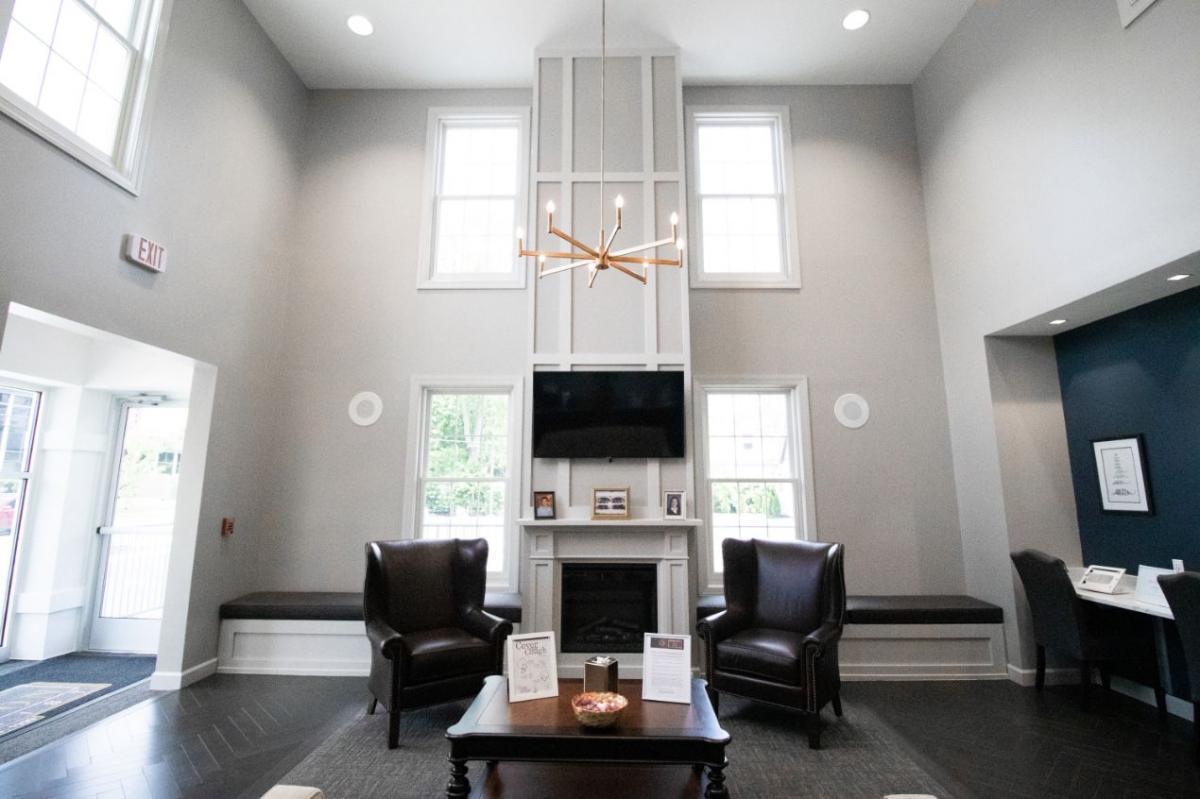 The image shows an interior view of a modern waiting room with a contemporary design featuring a neutral color palette, white walls, gray flooring, a fireplace, a large window, and multiple seating options including chairs and a couch.