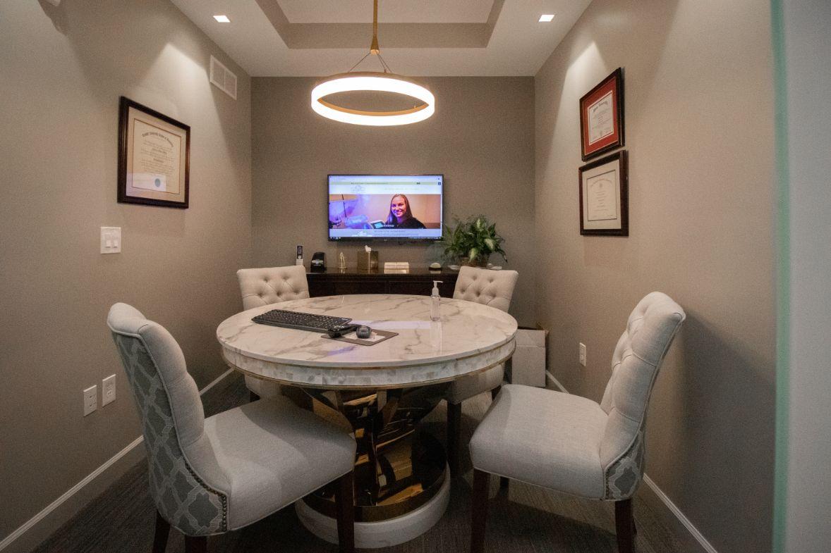 The image shows a well-lit room with a modern dining table and chairs, a television mounted on the wall, and framed certificates or awards displayed.