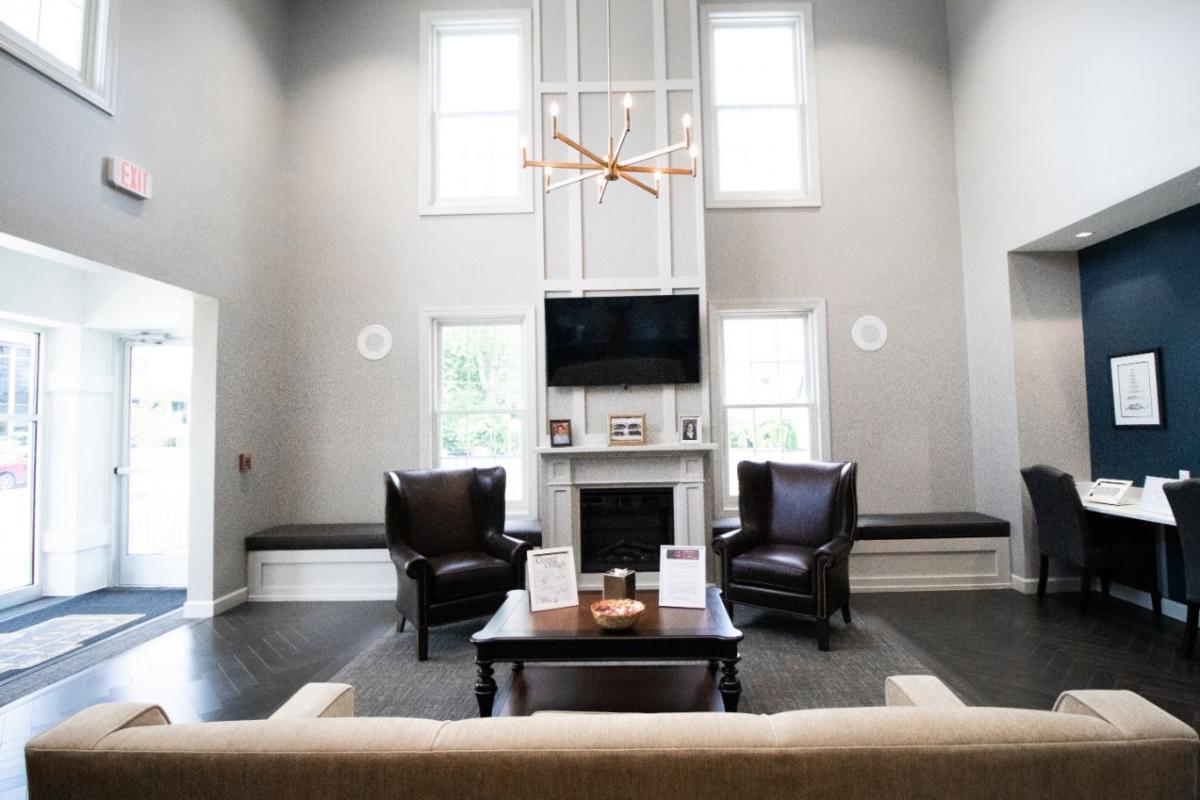 The image shows an interior space with a modern design, featuring a living room area with a sofa, chairs, and a coffee table, a dining area with a table and chairs, and a fireplace. The ceiling has exposed ductwork, and there s a large window allowing natural light to enter the room.