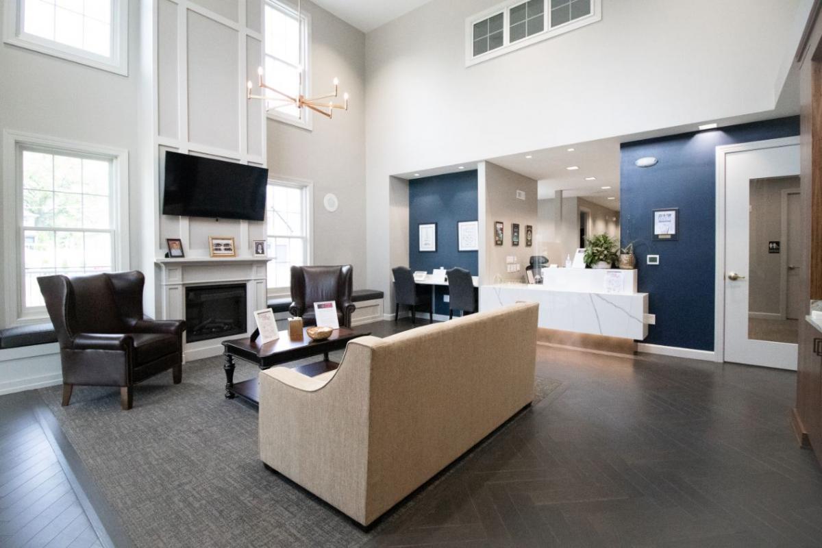 The image depicts a modern lobby with a contemporary design featuring a blue wall, white flooring, a comfortable seating area with a couch and chairs, a television mounted on the wall, and a reception desk with a computer monitor.