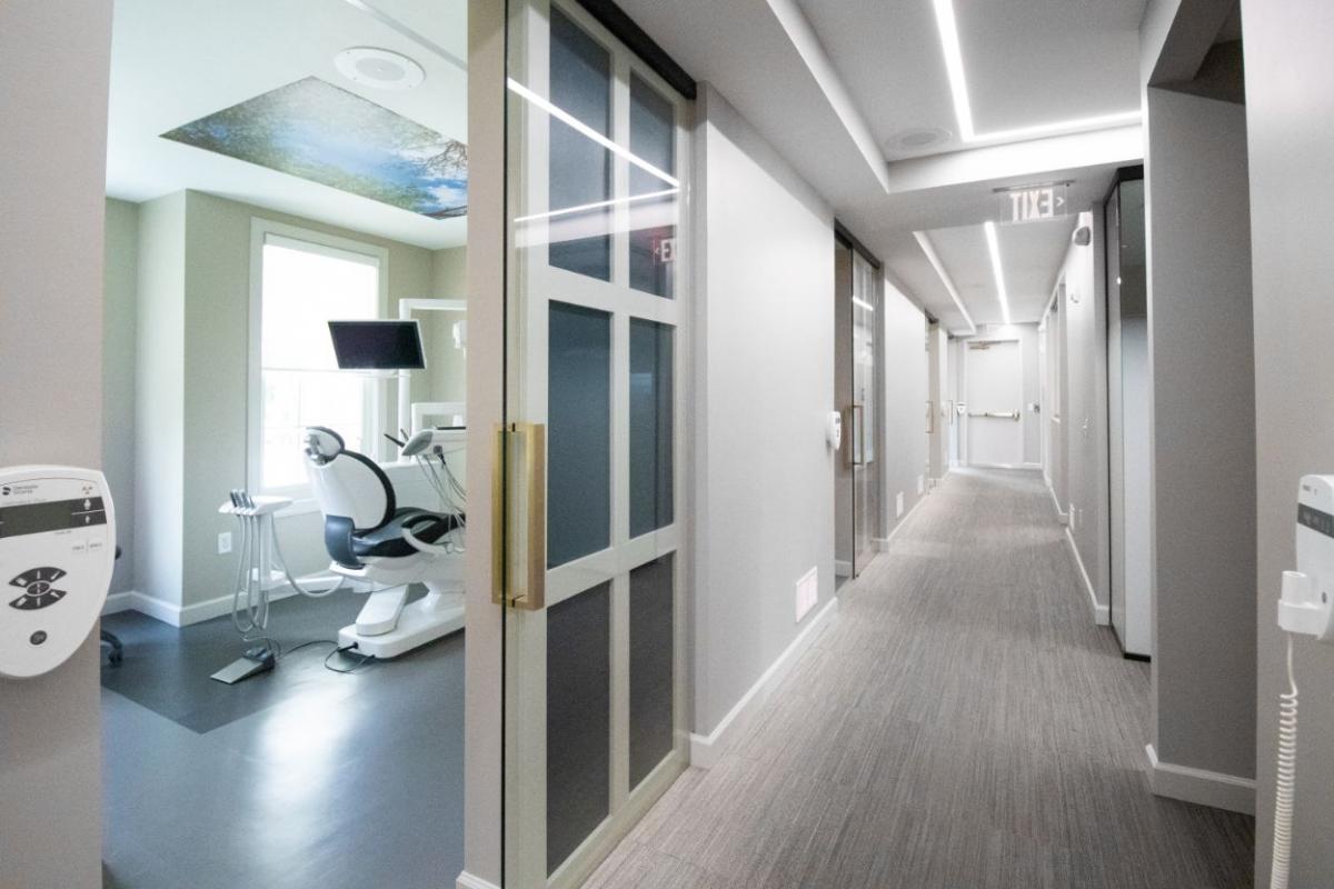The image depicts a modern, well-lit office hallway with a combination of interior design elements, including white walls, a sleek flooring, and contemporary furnishings such as chairs and a desk.