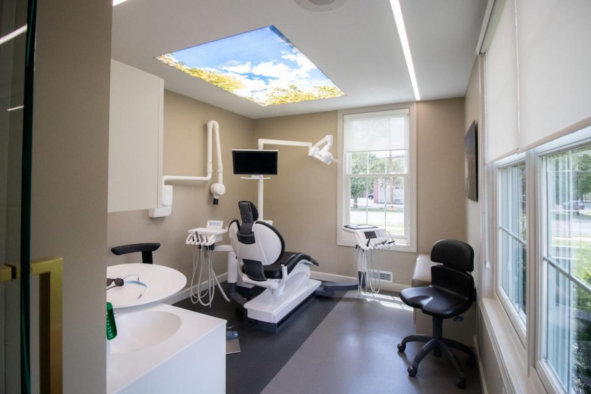 The image shows an interior space designed for dental services, featuring a clean and modern aesthetic with a ceiling-mounted skylight, a dental chair, and various equipment typically found in a dental office.