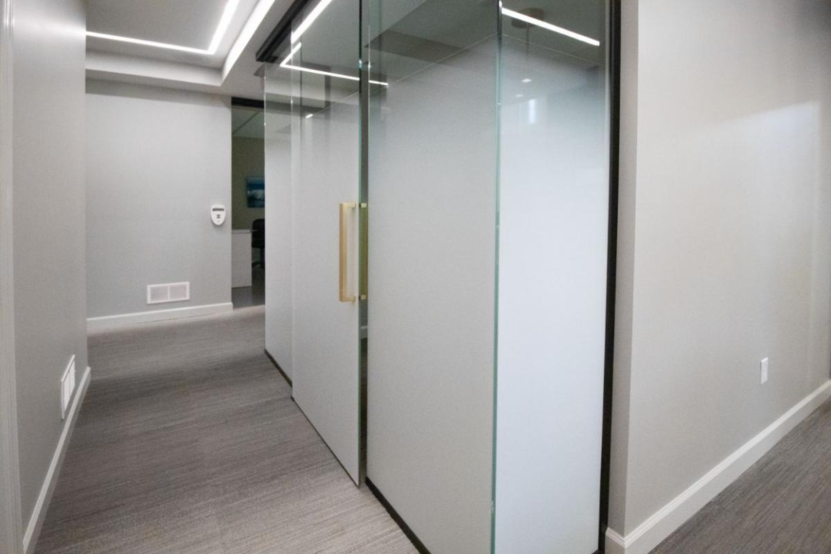 The image shows a modern office interior with glass walls and doors, a sleek design, and a hallway leading into a meeting room.