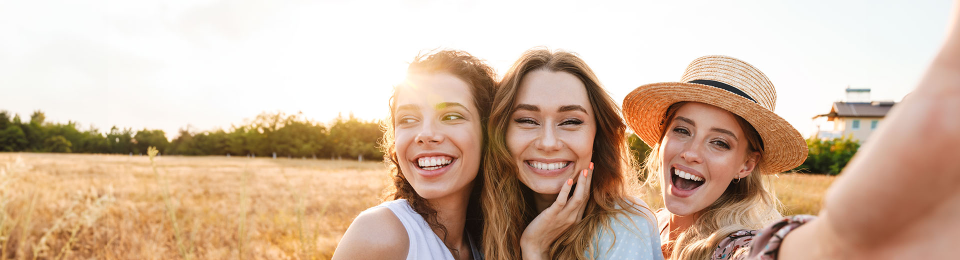 The image shows three smiling individuals taking a selfie outdoors during sunset.