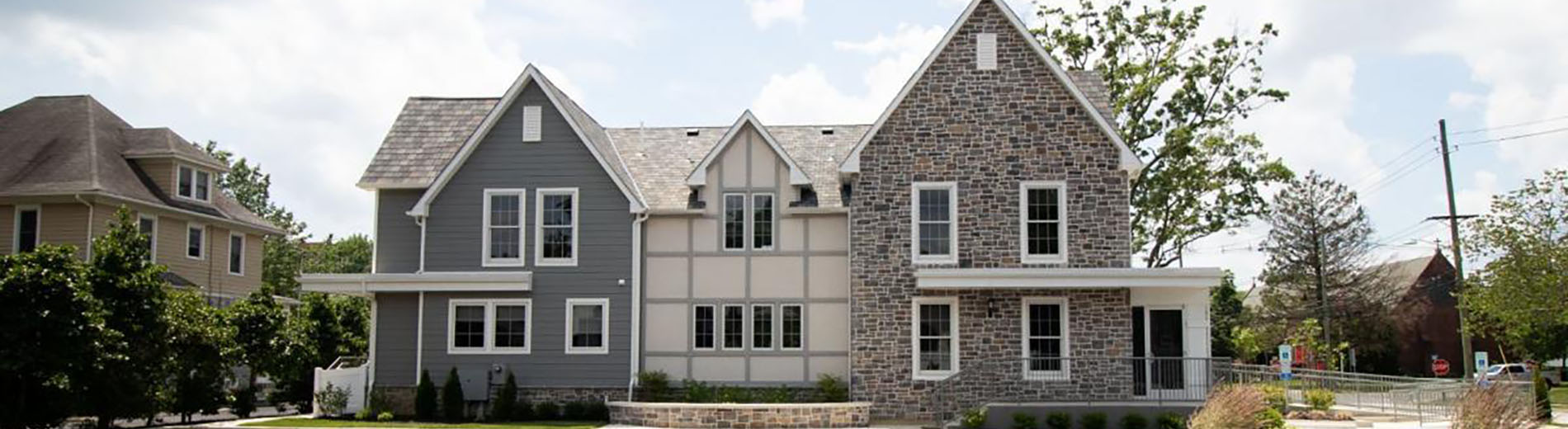 The image shows two-story houses with grey siding, white trim around windows and doors, and stone accents on the ground floor. There are trees in front of the houses, and a clear blue sky above.