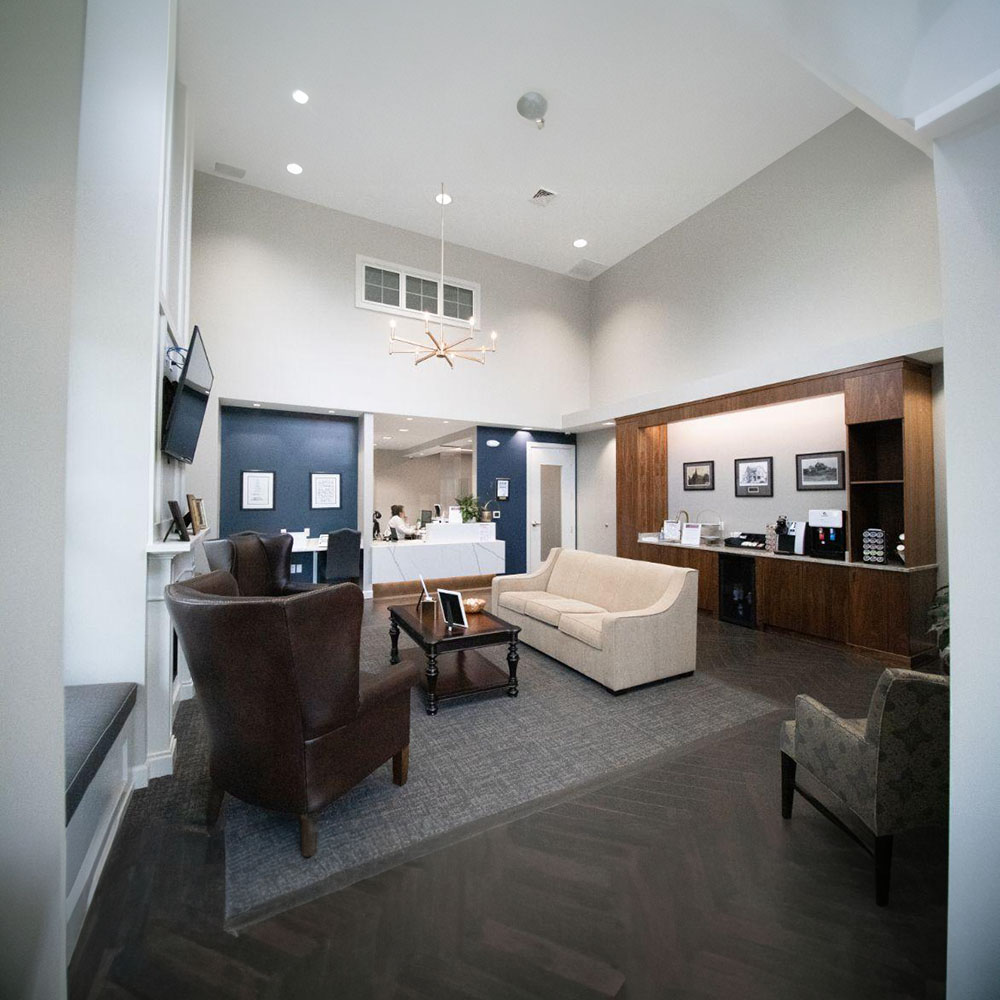 The image shows an interior space with a modern design, featuring a lobby area with a high ceiling, recessed lighting, and a clean, contemporary aesthetic. There are comfortable seating arrangements, including a couch and chairs, arranged around a central coffee table. A reception desk is visible in the background along with some wall decorations and artwork. The flooring appears to be carpeted, and there s a large window or glass partition that allows natural light into the space.
