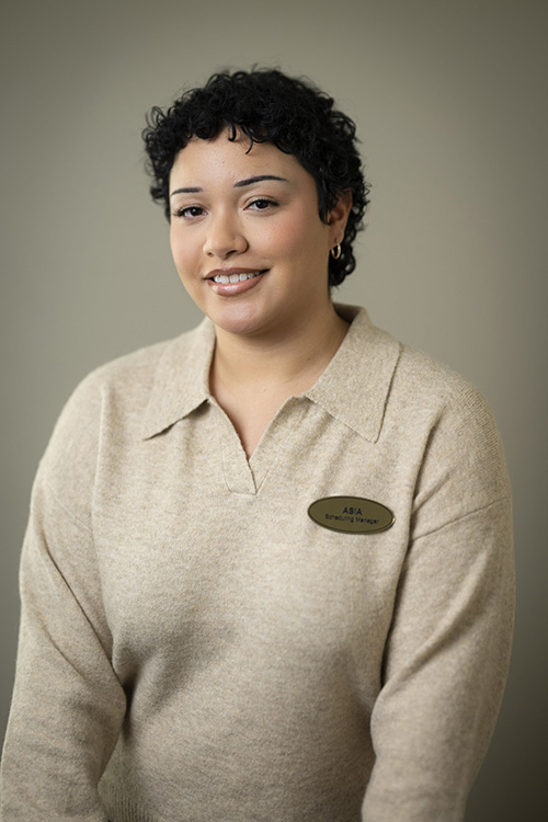 The image shows a person posing for a portrait with a slight smile, wearing a name tag and a beige sweater vest over a white shirt.