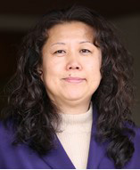 The image is a photograph of an adult woman with long hair, wearing a dark top and a light-colored blazer. She appears to be indoors, possibly in an office setting, and has a neutral expression on her face.