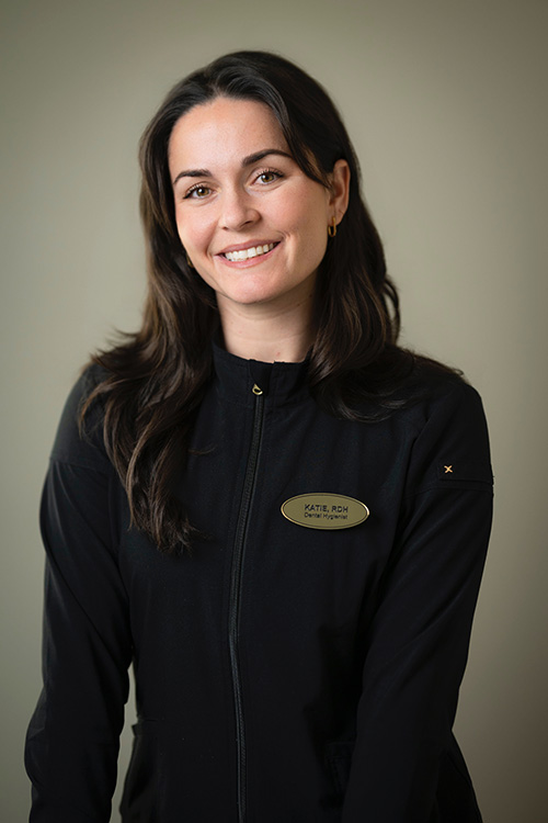 The image shows a woman posing for a portrait with a smiling expression, wearing a black jacket with visible name tags on a lanyard around her neck.