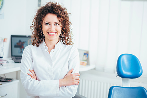 The image shows a woman with curly hair standing in an office environment, wearing a white shirt and smiling, leaning against a desk with a computer monitor and a chair behind her.