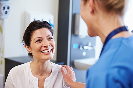 The image depicts two individuals in a medical setting, with one person seated and smiling at another standing person who appears to be a healthcare professional.