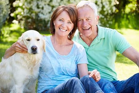 The image shows an older couple sitting on a grassy area with a golden retriever between them, both smiling and looking at the camera.