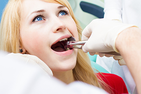 The image depicts a woman receiving dental care from a dentist, with her mouth open wide and a dental instrument being used on her teeth by a professional wearing gloves and a face mask.
