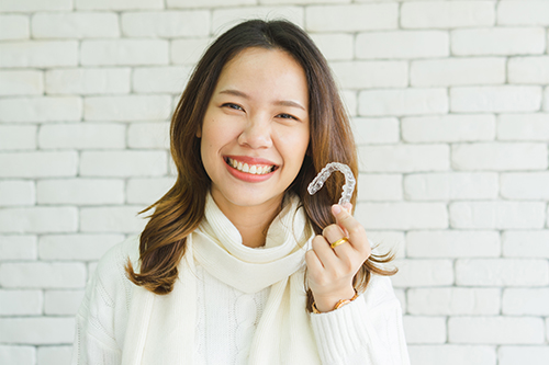 A smiling woman holding a white object with her right hand, posing against a brick wall background.