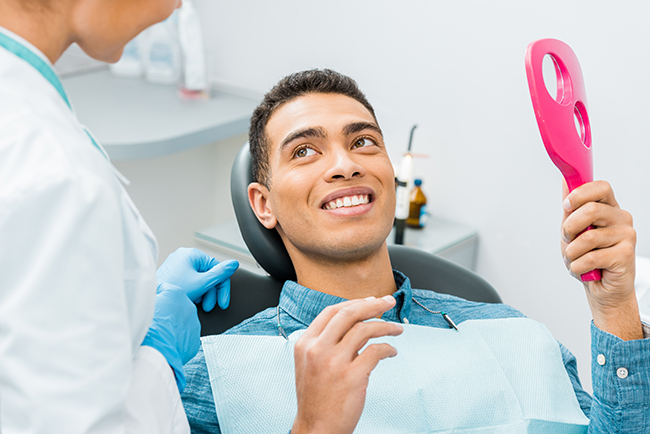 A dentist sitting in a dental chair with a smile, holding a pink toothbrush while receiving a dental cleaning service from another person.