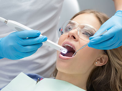 A woman wearing blue gloves and holding a device with a digital display near an open mouth being treated by dental professionals.